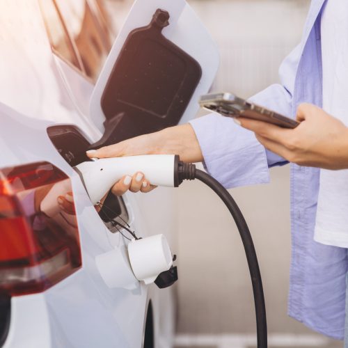 A woman charges her electric car while using a smartphone, reflecting modern eco-friendly technology and sustainable living in an urban environment.